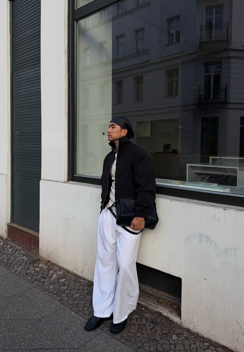 Jeune homme en veste noire et pantalon blanc appuyé contre un mur de bâtiment, tenant un sac noir et regardant vers la gauche.
