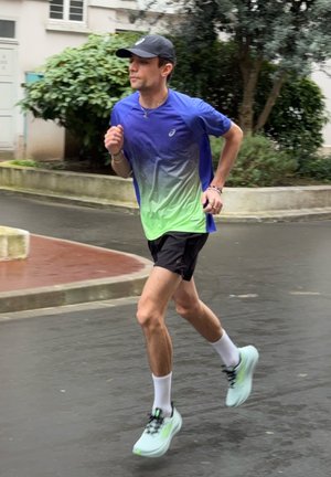 Hombre con camiseta azul verdosa para correr, pantalones cortos negros y gorra, trotando por una calle mojada cerca de vegetación y edificios.