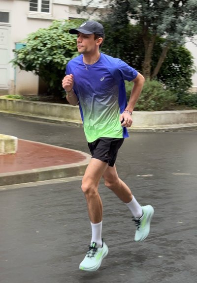 Hombre con camiseta azul verdosa para correr, pantalones cortos negros y gorra, trotando por una calle mojada cerca de vegetación y edificios.