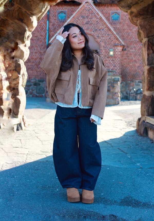 Young woman in a brown jacket and wide dark trousers stands outdoors framed by a stone archway with a brick building in the background.