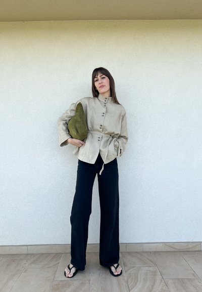 Chaqueta de lino beige claro con un cuello alto y botones, combinada con pantalones anchos negros y un bolso de mano verde, llevada con chanclas negras.