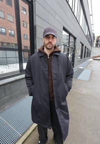 Άνδρας ντυμένος με μαύρο παλτό, καφέ φούτερ με κουκούλα και σκούφο στέκεται με τα χέρια στις τσέπες του σε αστικό πεζοδρόμιο κοντά σε μοντέρνα κτίρια.