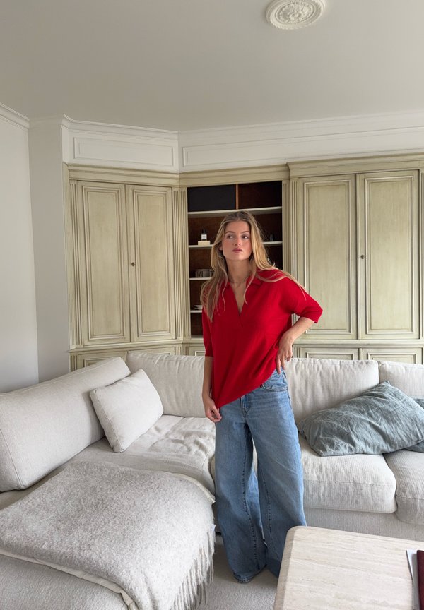 Blusa rossa a scollo a V con maniche a tre quarti abbinata a jeans a gamba larga di un blu chiaro. Divano dai toni neutri con cuscini in una stanza luminosa.