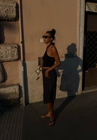 Fekete crop top és sötét bermuda rövidnadrág zsebekkel, mintás sál kiegészítőként. Napszemüveg és lapos szandál. Meleg esti fény.