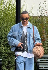 Jeansjacke über einer Lederweste, weißes Hemd und Jeans. Schwarze Sonnenbrille und ein gemusterter brauner Hut. Trägt eine braune Tasche mit einem Trageriemen.