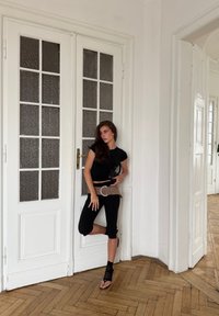 Camiseta ajustada negra y pantalones negros cortos, combinados con un cinturón ancho beige. El fondo presenta puertas blancas y suelo de madera.