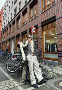 Person wearing a brown leather jacket with fur trim, a grey long-sleeve shirt, light-coloured trousers, and black shoes, leaning against a bicycle.