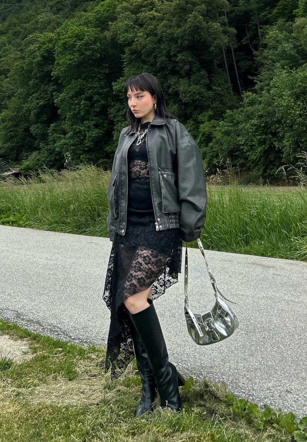 Robe en dentelle noire avec ourlet asymétrique, grande veste en cuir grise, bottes noires montantes et sac à main en argent avec une finition brillante.