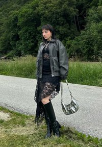 Robe en dentelle noire avec ourlet asymétrique, grande veste en cuir grise, bottes noires montantes et sac à main en argent avec une finition brillante.