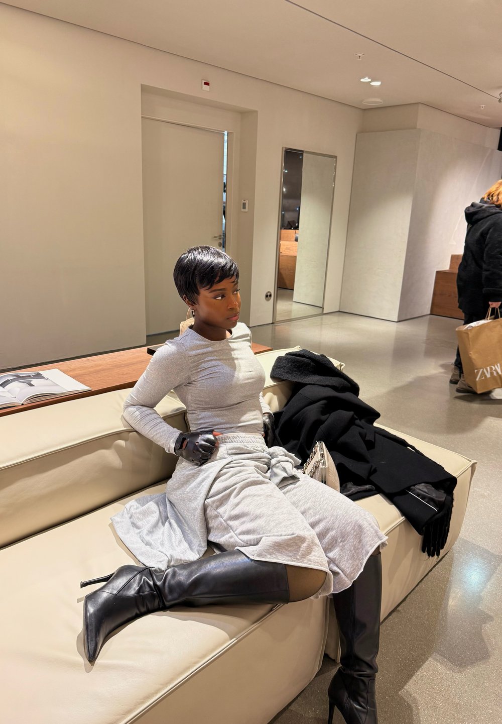 Femme portant un haut gris à manches longues, une jupe grise, des gants noirs et des bottes noires hautes jusqu'aux genoux, assise sur un canapé beige dans une pièce moderne.