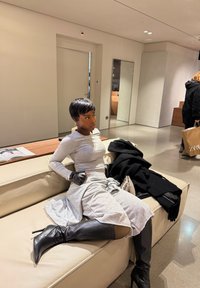 Femme portant un haut gris à manches longues, une jupe grise, des gants noirs et des bottes noires hautes jusqu'aux genoux, assise sur un canapé beige dans une pièce moderne.