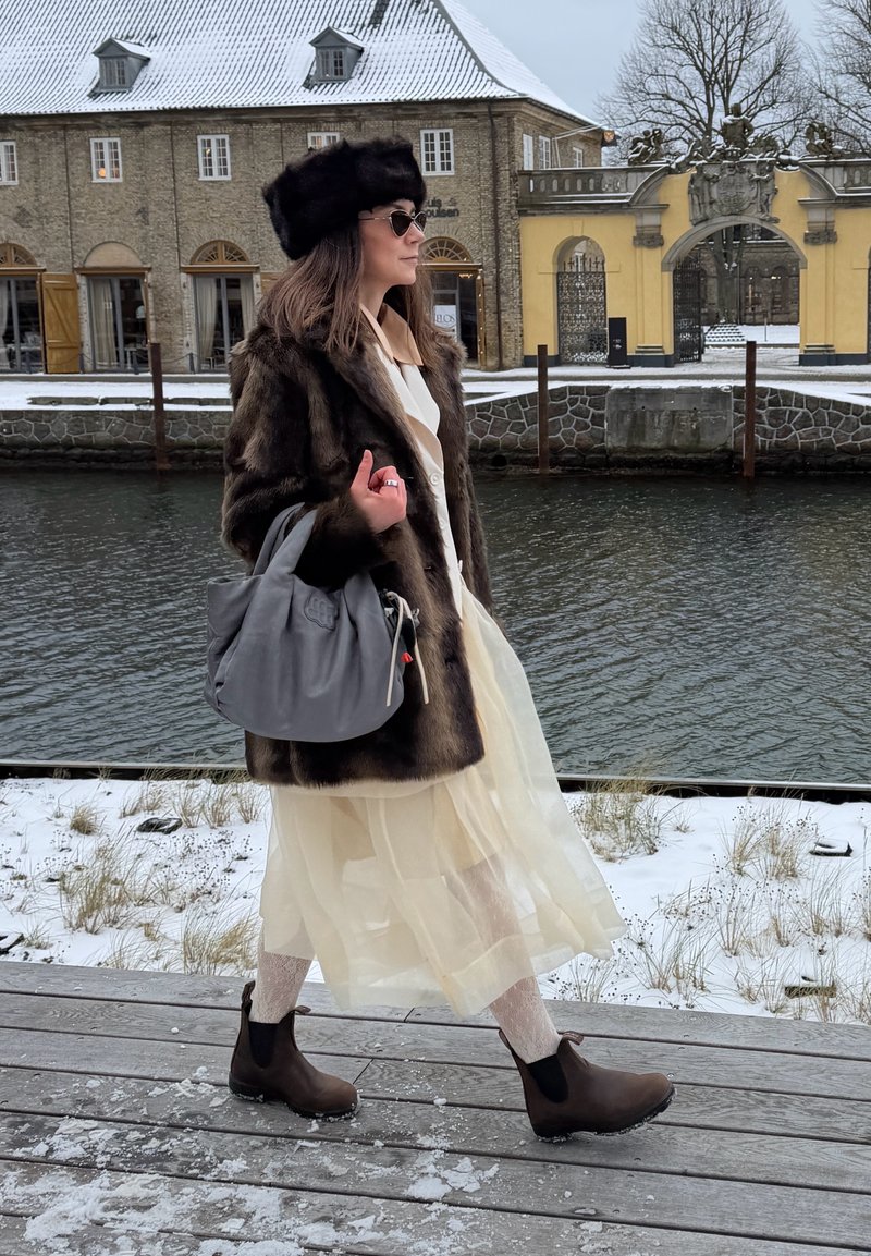 Donna con cappotto di pelliccia, cappello nero e occhiali da sole che cammina su un molo di legno innevato accanto al fiume, con edifici storici sullo sfondo.