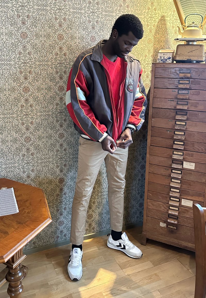 Νεαρός άνδρας με κόκκινο πουλόβερ και γκρι μπουφάν με κόκκινες και λευκές ρίγες, κλείνει το φερμουάρ του μπουφάν, στέκεται δίπλα σε ξύλινη συρταριέρα σε δωμάτιο με μοτίβο.