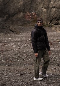 Černá péřová bunda s kapucí a předním zipem, zelené kalhoty a b�ílé tenisky. Sluneční brýle a hnědá čepice s logem. Skalnatý terén v pozadí.