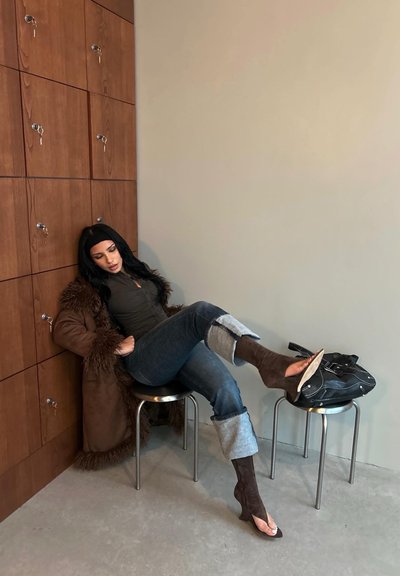 Mujer con un abrigo con ribete de piel y vaqueros remangados sentada en un taburete, con una pierna apoyada en otro taburete, sosteniendo un bolso negro.