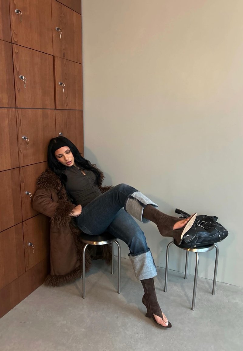 Femme portant un manteau bordé de fourrure et un jean retroussé, assise sur un tabouret, une jambe reposant sur un autre tabouret, tenant un sac noir.