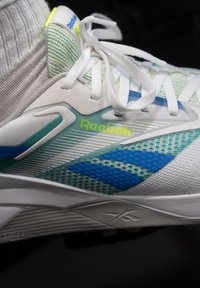 Hvid atletiksko med mesh-øvre, der har grønne og blå accenter, strukturerede overlæg og snørebånd. Reebok-logoet er tydeligt vist.