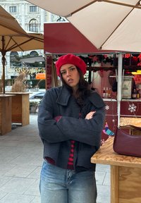 Jeune femme portant un béret rouge et une veste gris foncé, debout les bras croisés près d'une table en bois et des étals de marché en plein air.