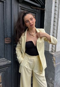 Ανοιχτό κίτρινο κοστούμι με λεπτές ρίγες, διαμορφωμένο σακάκι και παντελόνια σε φαρδιά γραμμή, συνδυασμένα με ένα μαύρο strapless τοπ και περιδέραιο με χρυσές λεπτομέρειες.