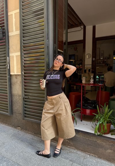 Camiseta negra ajustada con la palabra "DELICIOUS" impresa, pantalones cortos de pierna ancha en color beige y zapatos planos negros. Planta verde en una maceta y una mesa circular roja en el fondo.