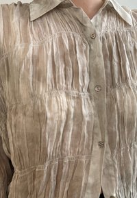 Světle barevná, texturovaná košile s knoflíky v neutrálních odstínech se svislými sklady a jemným límcem. Obsahuje knoflíky na přední straně a nařasené detaily.