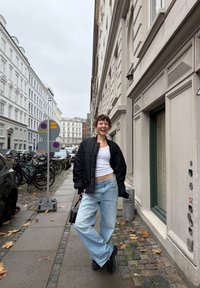 Černá bomber bunda, bílý crop top, světle modré široké džíny. Černé platformové boty. Ulice s dlažebními kostkami, jízdními koly a fasádami budov.