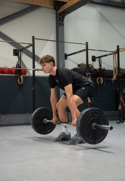Joven en una camiseta atlética negra y pantalones cortos, levantando una barra con grandes pesas negras en un gimnasio con suelo gris y características industriales.