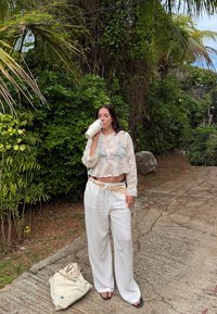 Haut court en dentelle blanche à manches longues, associé à un pantalon en lin blanc ample et une ceinture beige. Tenant une tasse de café, debout sur un chemin.