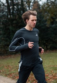 Muž s hnědými vlasy v černém sportovním oblečení běží venku po cestě, v pozadí jsou tráva a stromy.