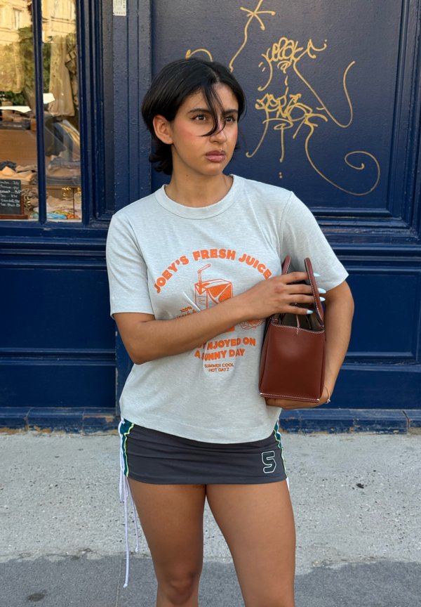 Jonge vrouw met kort zwart haar, draagt een lichtgrijs T-shirt met grafische opdruk en een donkere miniskirt, houdt een bruine handtas vast bij een blauwe muur met gouden graffiti.