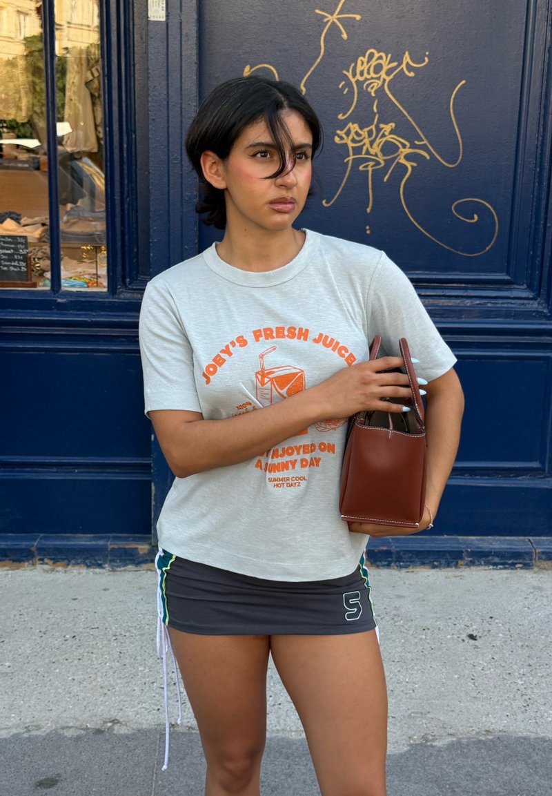 Jeune femme aux cheveux noirs courts portant un t-shirt graphique gris clair et une mini-jupe foncée, tenant un sac à main marron près d’un mur bleu avec un graffiti doré.