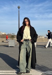 Femme portant des lunettes, un long manteau noir, une chemise rayée et un jean large, debout dans une rue avec des personnes et des bâtiments en arrière-plan.
