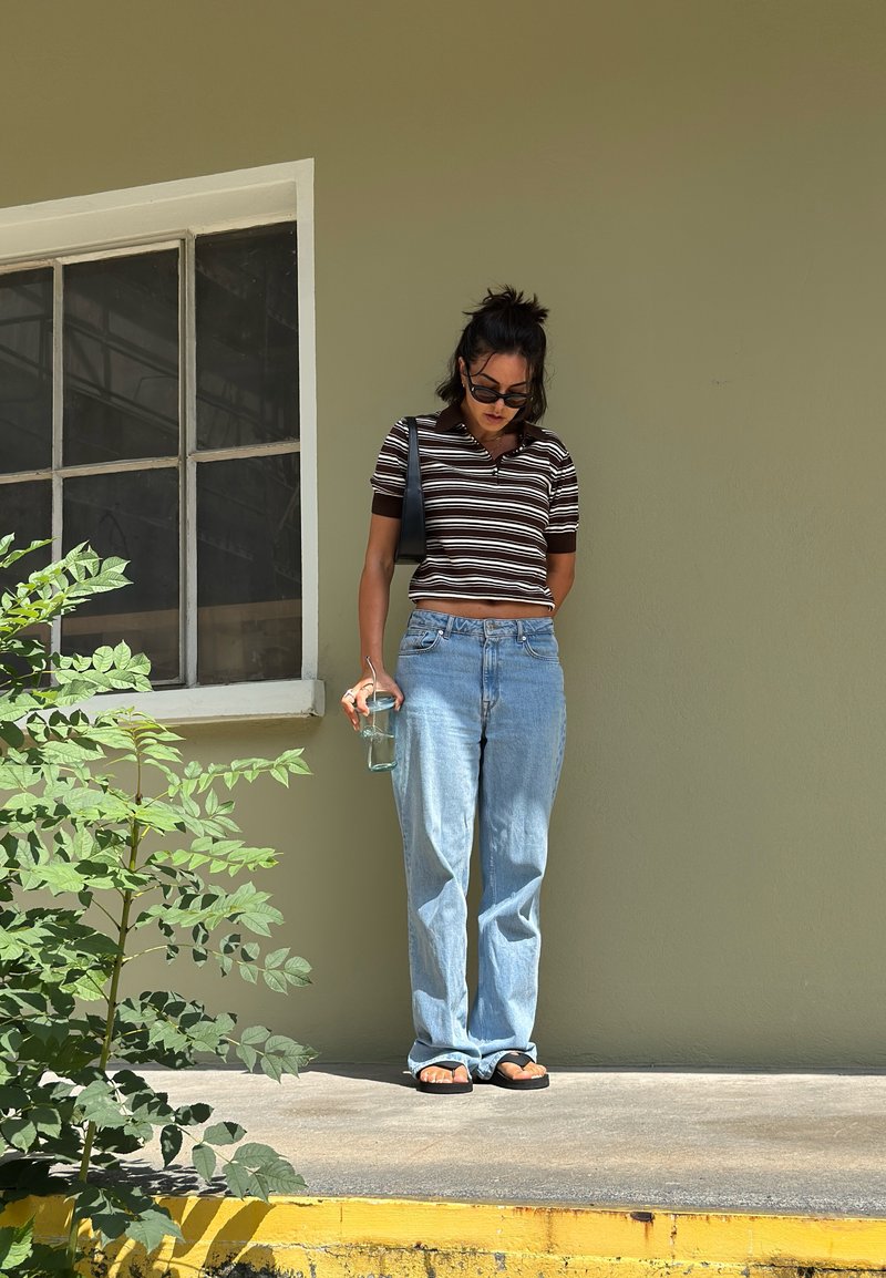 Top corto a strisce marrone e bianco, jeans larghi blu chiaro, sandali neri; tiene in mano un bicchiere trasparente, in piedi su una superficie grigia vicino a una pianta.