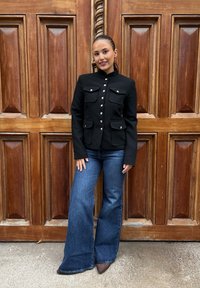 Svart skreddersydd blazer med fire frontlommer og sølvknapper, kombinert med vide blå denimjeans og brune hælede sko. Bakgrunn av trepanel.