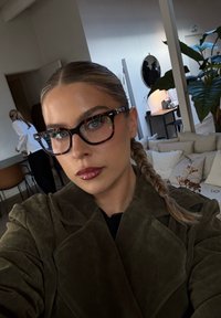 Femme aux cheveux tressés, portant des lunettes écaille de tortue et un manteau vert foncé, prenant un selfie dans une pièce moderne avec un canapé, des plantes et des personnes en arrière-plan.