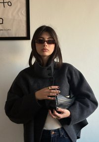 Manteau en laine gris surdimensionné avec un col montant, des boutons noirs et un sac à main texturé. Le modèle a les cheveux foncés et porte des lunettes de soleil rectangulaires.