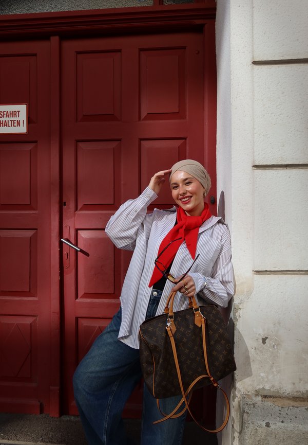 Una mujer sonriente con un pañuelo beige en la cabeza, una bufanda roja, una camisa de rayas y vaqueros posa junto a una puerta roja. Sostiene unas gafas de sol y un bolso grande de color marrón.