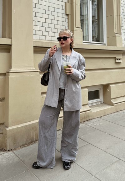 Blazer de corte oversize en gris claro y pantalones de pierna ancha a juego con una textura arrugada. Bolso negro y gafas de sol. Sosteniendo una bebida verde.