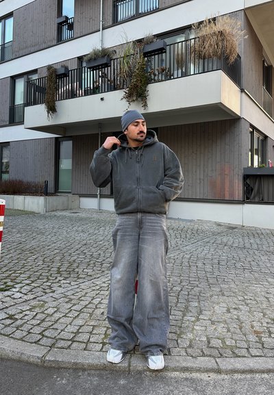 Sudadera gris con cremallera, capucha grande y bolsillos frontales, combinada con unos pantalones vaqueros grises holgados y zapatillas blancas sobre un fondo de piedra pavimentada.