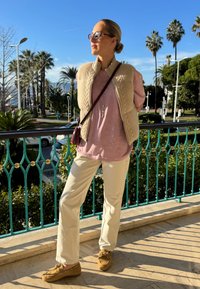 Femme portant des lunettes de soleil, un gilet beige, une chemise rose, un pantalon crème et des chaussures marron, debout sur un balcon avec des palmiers et un ciel bleu en arrière-plan.
