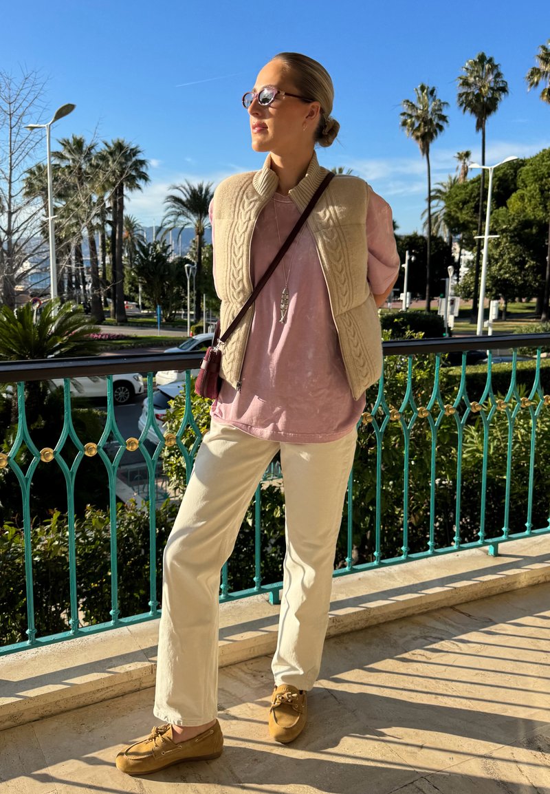 Femme portant des lunettes de soleil, un gilet beige, une chemise rose, un pantalon crème et des chaussures marron, se tient sur un balcon avec des palmiers et un ciel bleu en arrière-plan.