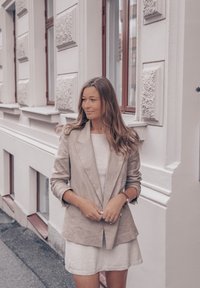 Lichtgrijze linnen blazer met een gestructureerd ontwerp, gedragen over een textuur witte top en een witte jurk, gecombineerd met een zilveren armband.