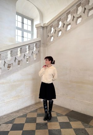 Mujer con suéter crema, falda negra y botas altas de pie junto a una escalera de piedra ornamentada con una gran ventana en un pasillo luminoso con azulejos.