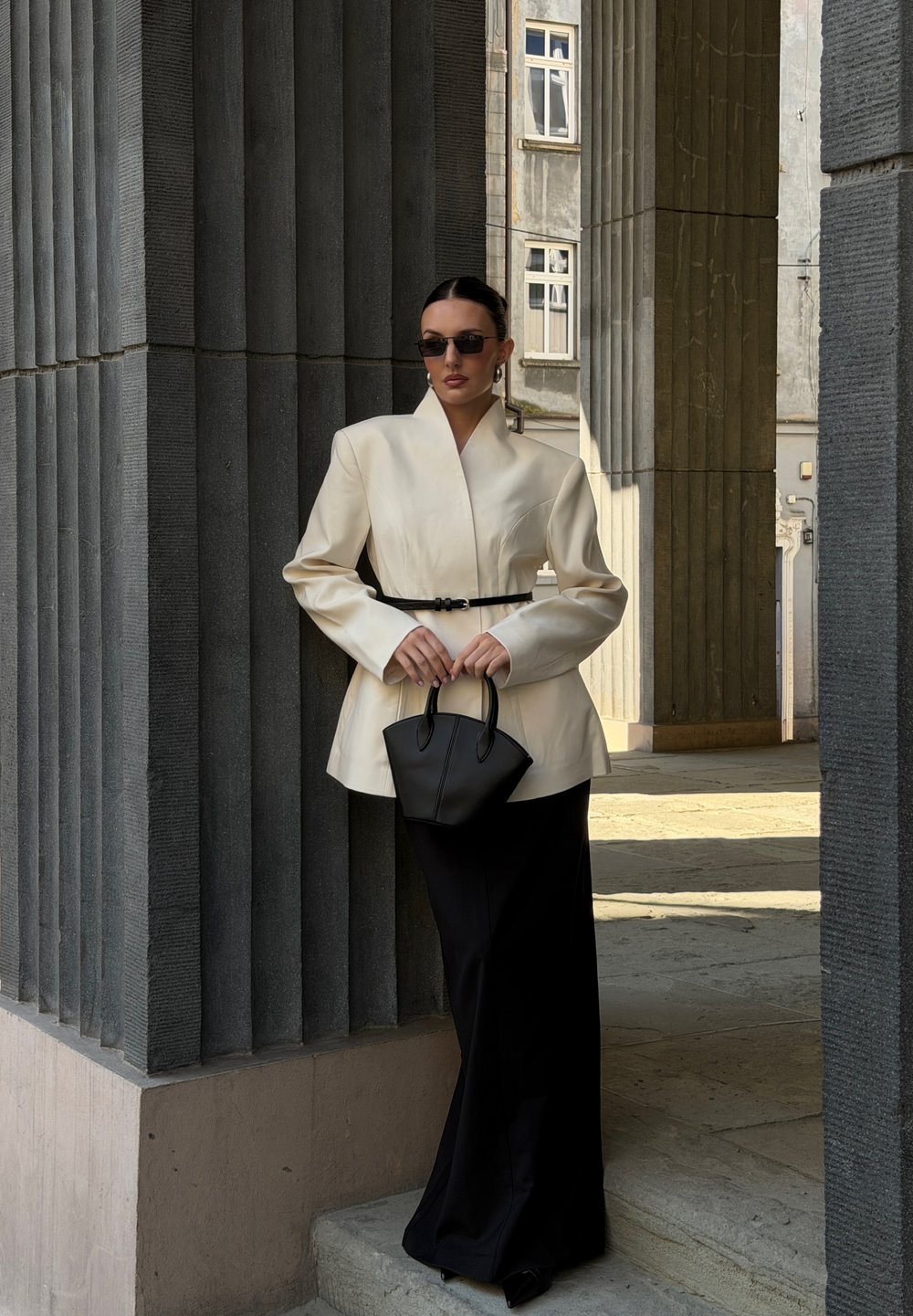 Beige gestructureerde blazer met een ceintuur in de taille, gecombineerd met zwarte wijde pantalons. Houdt een kleine zwarte handtas van gestructureerd leer vast.