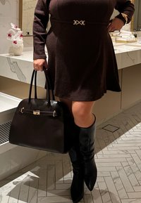 Brūnas rievotas kleitas ar zelta jostas detaļu, kopā ar melnu ādas somiņu un melniem ceļgala augstiem zābakiem uz rakstaina grīdas.