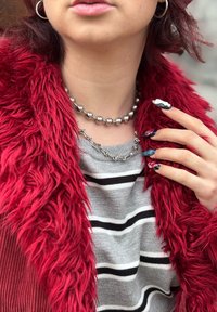 Vêtement extérieur en fausse fourrure rouge, pull rayé gris, deux colliers en métal et ongles longs avec des motifs complexes. Apparence texturée et superposée.