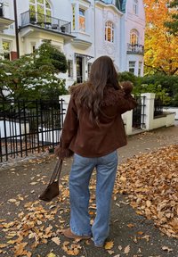 Jaqueta de camurça castanha com colarinho e punhos de pele, combinada com jeans claros de boca larga. A carregar uma pequena mala castanha, rodeada de folhas de outono.