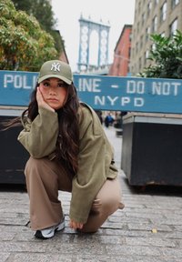 Veste oversize vert olive, pantalon large marron et baskets grises. Porte une casquette verte avec un logo blanc, posé devant un panneau de police bleu.