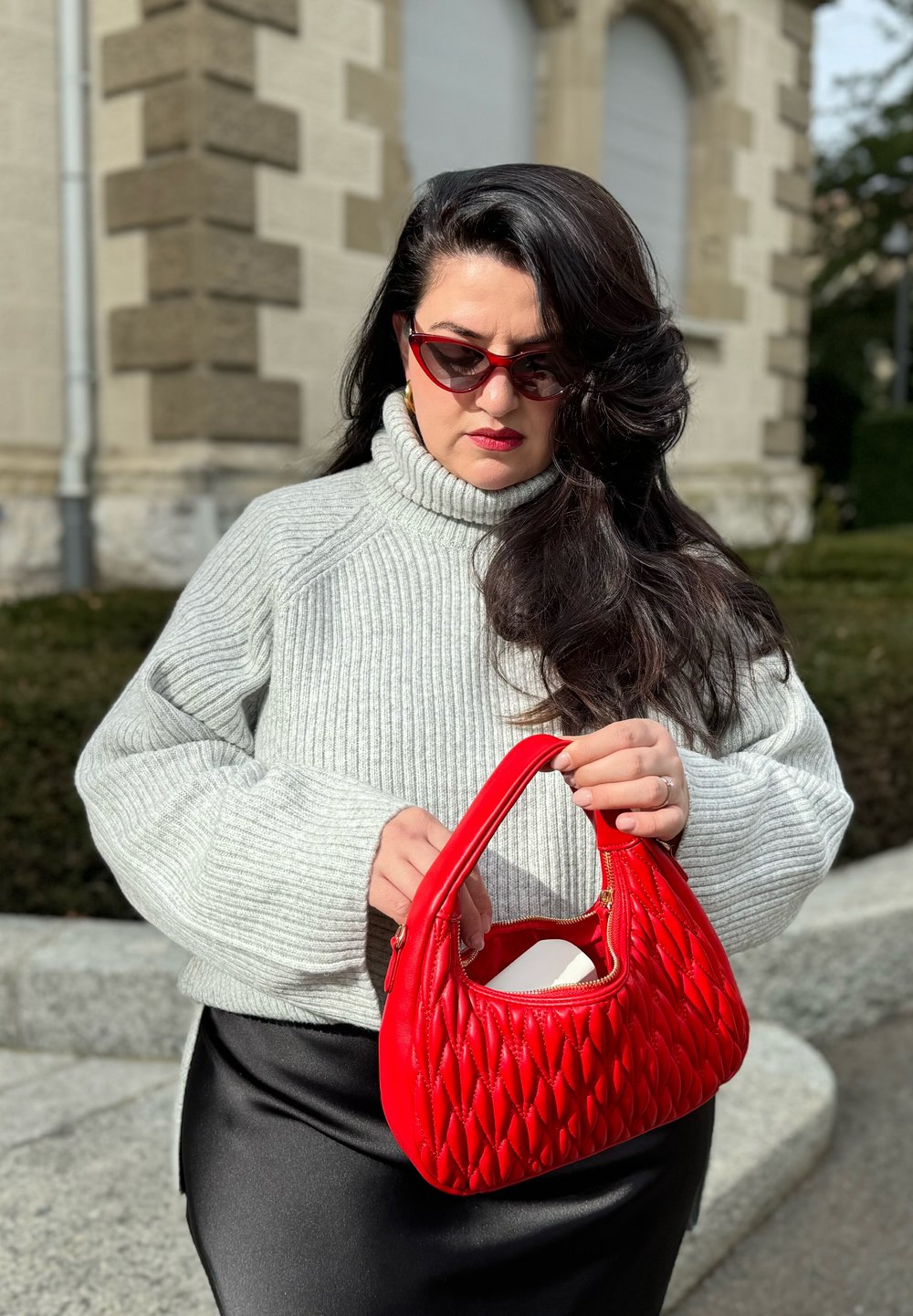 Κόκκινη κεντημένη τσάντα με δομημένο σχήμα και καμπύλη λαβή, συνδυασμένη με γκρι ριχτό πουλόβερ με γιακά και μαύρη δερμάτινη φούστα.