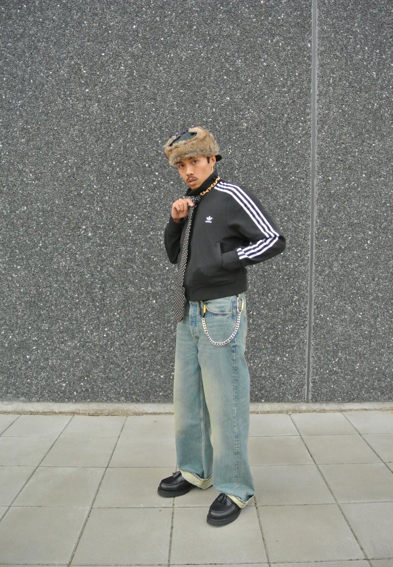 Μαύρο μπουφάν με φερμουάρ και λευκές ρίγες, τζιν παντελόνι με ξεθωριασμένο φινίρισμα, μαύρα παπούτσια και καπέλο με γούνα. Περιλαμβάνει αξεσουάρ αλυσίδας και γραβάτα με σχέδιο.